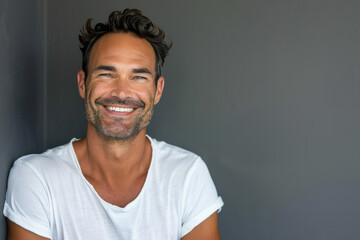 A man in a white shirt is smiling and leaning against a wall