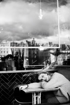 Relaxed Woman Enjoying City View in Cafe. A serene woman sits at a cafe table, gazing out the window at the cityscape. Her peaceful demeanor reflects contentment as she savors the urban scenery.