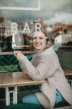 Smiling Woman Enjoying Coffee in Cafe. A cheerful woman sits at a cafe table, holding a cup of coffee with a visible bar sign in the background. Warmth and relaxation exude from the cozy atmosphere