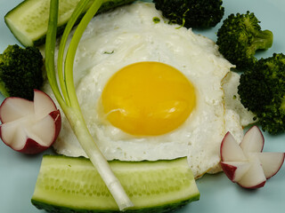 Fried egg, boiled broccoli, radishes, fresh cucumbers and green onion are on a pale blue plate, healthy breakfast     
        