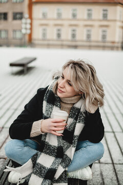 A woman sits on a bench, wrapped in a scarf, enjoying a drink. The serene park setting evokes a sense of relaxation and tranquility. Cozy Moment in the Park