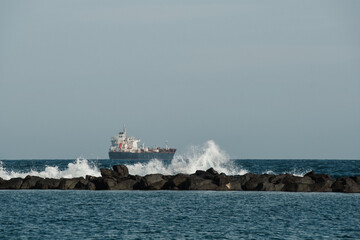 Waves crashing against a breakwater in the ocean with a ship at anchor in the background. Waves brake and splash against the rock. Big ship in the distance with a copy space for a free text