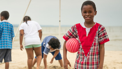 Portrait of a black boy on a beach, holding a ball in his arm.