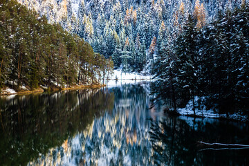 Paisagem de um lago na floresta, com árvores e neve, onde se vê o reflexo de tudo na água