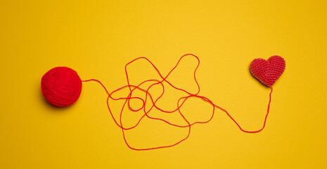 A ball of thread and a knitted red heart on a yellow background, top view