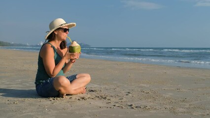 Cheerful young woman in straw hat enjoying summer vacation and drinking coconut water on the beach. Copy space