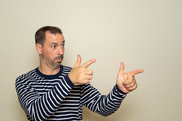 Hispanic man with beard with two hands arms sign gesture pointing aside, isolated beige wall background. Feelings, signs and facial expression symbols of positive human emotions
