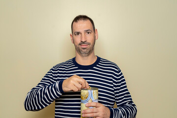 A Hispanic man with a beard wearing a striped sweater putting a euro coin into a metal piggy bank, trying to save so he can travel on vacation. Isolated on beige studio background.