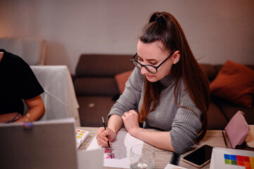 Two artists engage in a collaborative discussion over a watercolor palette, with one offering guidance as they navigate the hues together.