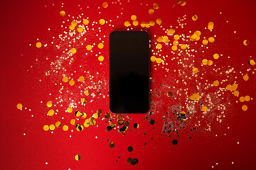 New mobile phone lies on a red background among silver sequins and round gold confetti. Phone with...