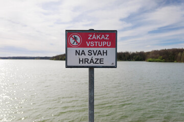 Warning sign with text - Do not enter on the dam embankment (translated from Czech). Prohibition, bad and interdiction. Water reservoir in the background. Shallow focus.