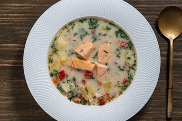 Fresh creamy salmon fish soup with potatoes, carrots, peppers and onions in a ceramic plate on a wooden table. A delicious dinner consists of fish soup with salmon