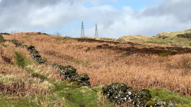 Dry stone wall with in distance overhead electric power lines and poles across brown hills of the Scottish Highlands, delivering power en energy under a dark storm sky with bright clouds
