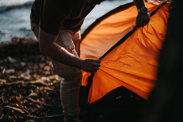A close-up view of friends working together to set up an orange tent near a lake, enjoying their...
