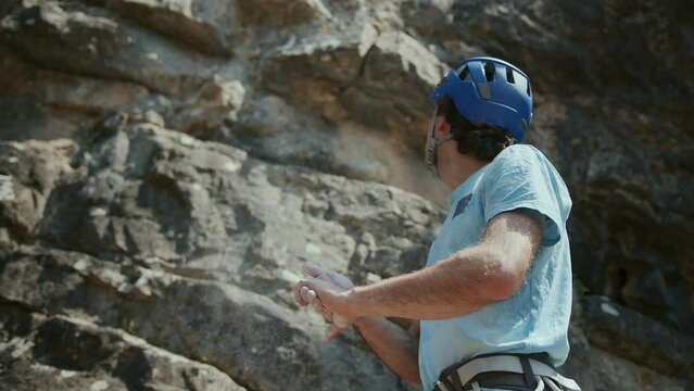 A male rock climber is going to climb a rock. He claps his hands and the chalk dust comes from them. He wears a blue helmet. Concept of overcoming, success and achievement.