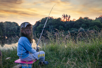 Young boy sitting with fishing rod on the edge of a high riverbank at sunset with vibrant colors on...