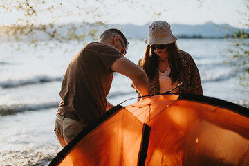 A young man and woman work together to pitch an orange tent by a lake, capturing a joyful moment of...