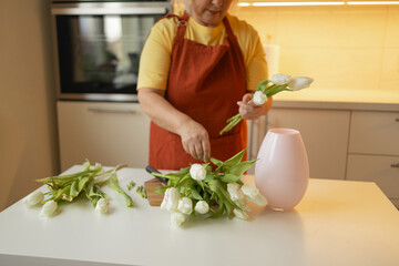 Happy Caucasian 30s woman taking care of cut fresh bouquet of tulips in kitchen. Housewife hands of woman cutting flower on wooden board and putting fresh tulips into the vase.