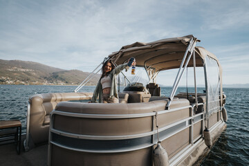 A happy, youthful woman raises her drink in celebration while standing on a pontoon boat, surrounded by a scenic lake view. © qunica.com