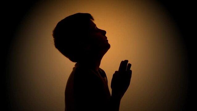 A young boy is praying to God above with his hands in a shadow with light for a Christianity faith concept or bible school idea.