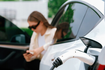 Young woman use smartphone to pay for electricity at public EV car charging station green city...