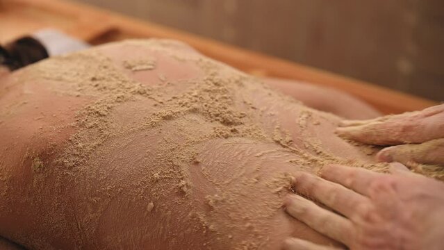 Relaxed female client getting back massage with scudding sand in resort spa salon. Closeup of young woman receiving body massage in luxury wellness center. Body relaxation and skin care, slow motion.