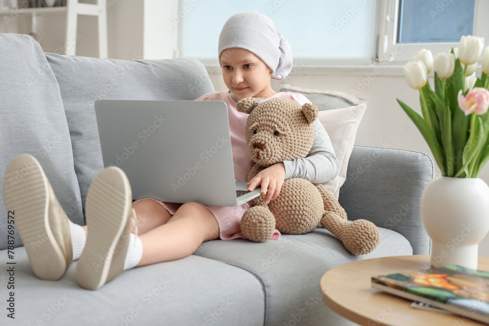 Wall mural Little girl after chemotherapy with toy bear using laptop on sofa at home. International Childhood Cancer Day