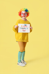 Funny little girl in clown wig with card on yellow background. April Fools' Day celebration