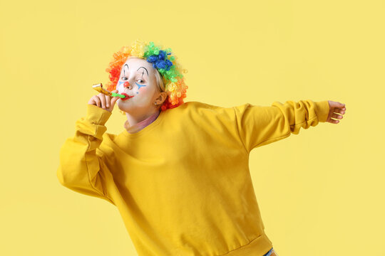 Funny little girl in clown wig with party blower on yellow background. April Fools' Day celebration