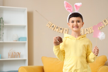 Cute little boy in bunny ears headband holding chocolate egg and decor for Easter at home