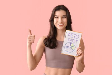 Happy smiling young woman holding notebook with keto diet chart and showing thumb-up on pink...