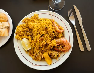 Poster Crumbly paella with seafood, decorated with large tiger shrimps, complemented with quarter of lemon, and glass with beverage. Festive dish serving, eating out, concept of tasty, healthy, healthy © JackF