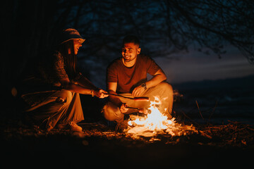 Two happy friends camping by a lake, preparing dinner over a cozy campfire under the evening sky.