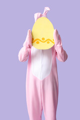 Young man in bunny costume with paper Easter egg on lilac background