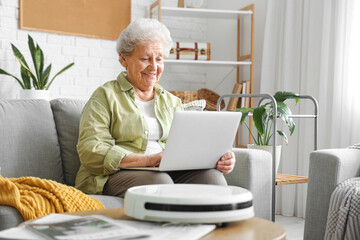 Senior woman with robot vacuum cleaner and laptop at home