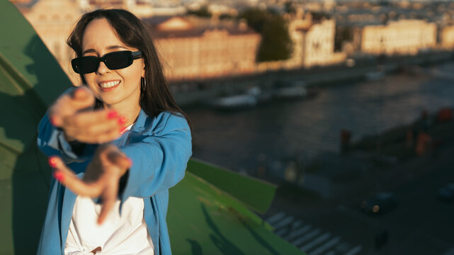 Happy young woman wearing sunglasses and blue blazer standing on rooftop of city building, smiling, puts his hands out
