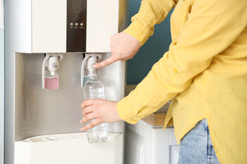 Woman filling plastic bottle with filtered water from cooler near blue wall