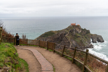 Panoramic view of the hermitage of San Juan de Gaztelugatxe in the middle of the sea on the touristy Basque coast and a Japanese tourist photographing the landscape on a cloudy day.