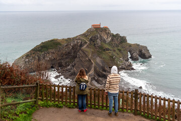 Couple of tourists with their backs to San Juan de Gaztelugatxe taking pictures of the hermitage surrounded by the sea on the touristy Basque coast.