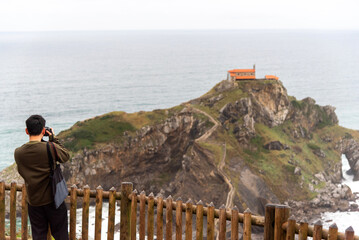 View of the hermitage of San Juan de Gaztelugatxe on the touristy Basque coast and a Japanese tourist with his back turned and his professional camera photographing the landscape.