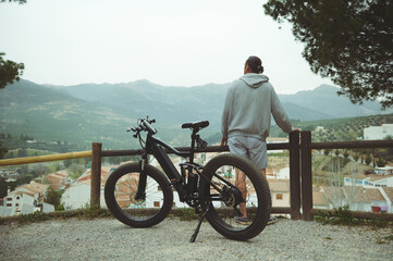 Rear view of a young active man cyclist standing near his electric battery powered bike, admiring...