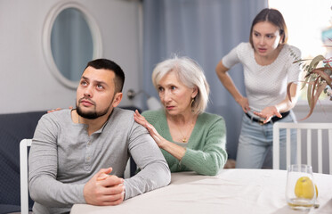 Portrait of displeased young adult man sitting at home ignoring his wife berating him, while her mother embracing and soothing him