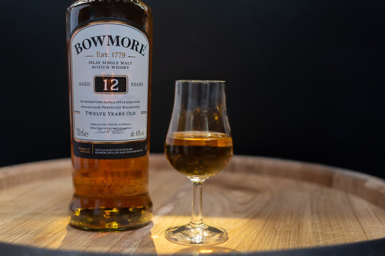 Bottle of Islay single malt scotch whisky Bowmore 12 years aged with whisky glass close-up on oak cask in soft light on blurred black background. Ukraine, Zhytomyr, April, 17, 2024