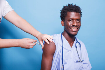 Male medical nurse wearing blue scrubs getting vaccine injection with syringe in studio. African American physician with stethoscope, getting vaccinated for coronavirus immunity. - obrazy, fototapety, plakaty