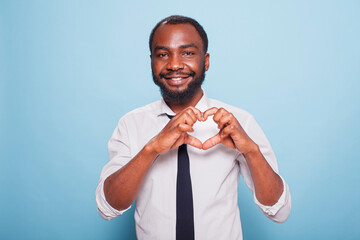 Smiling african american influencer making heart shape fingers to show care for supporting fans. Joyful male blogger in white shirt and black tie doing the hand gesture for love on his chest.