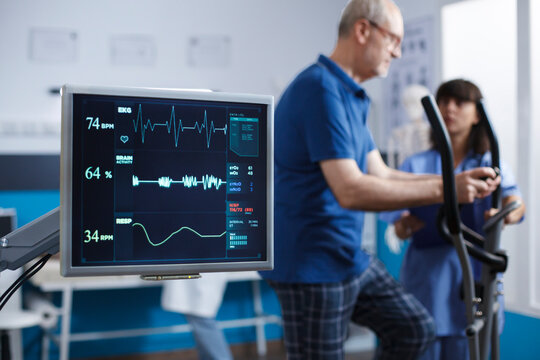 Close-up of heart rate monitor taking a pulse of senior patient during physical rehabilitation activity. Screen showing vital signs of elderly man while he uses stationary bicycle for physiotherapy.