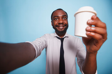 Black man having a video call while holding up cup of coffee against isolated background. Smiling african american dressed in white shirt making a selfie photo with beverage container in his hand.
