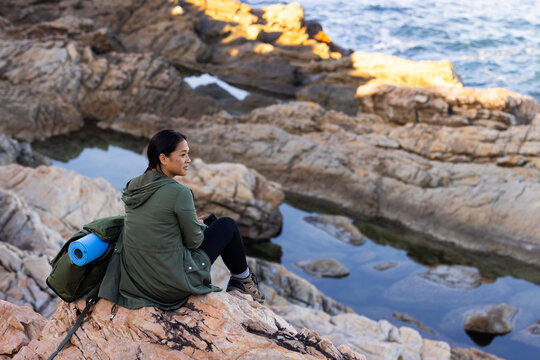 Biracial female hiker sitting on rocky shore, looking at sea, surrounded by the serene beauty of nat