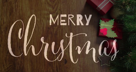 Merry Christmas written in cursive on wooden surface, surrounded by holiday decor