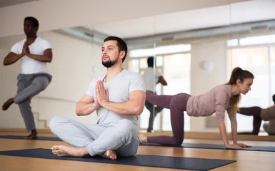Rugzak Caucasian man sitting on mat in lotus pose during group yoga training in gym. © JackF